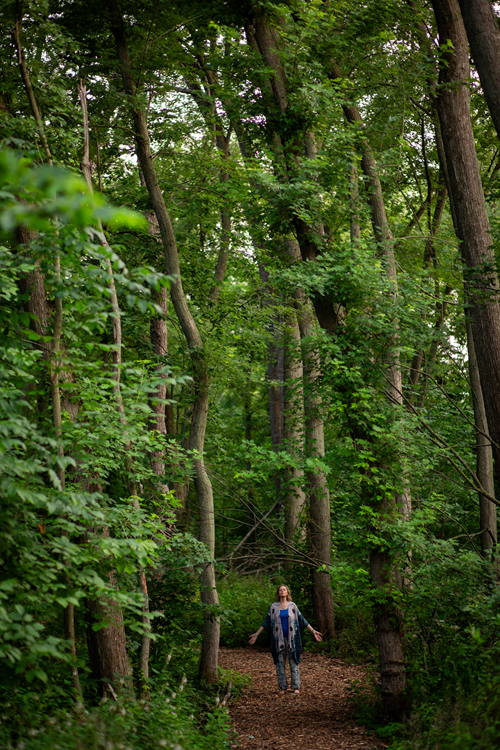 Picture of a female standing arms wide open looking 
 up to the sky in the middle of a path in the forest
